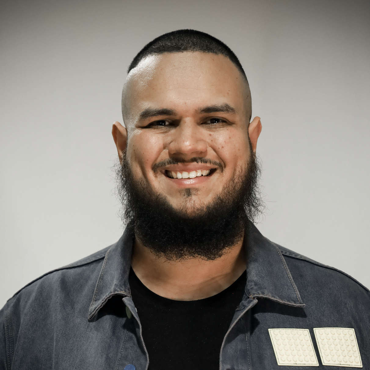 A man with a closely shaved head and a full beard is smiling at the camera. He is wearing a dark denim jacket over a black shirt. The background is plain and neutral, allowing the focus to be on his face and expression. He exudes warmth and friendliness with his broad smile.